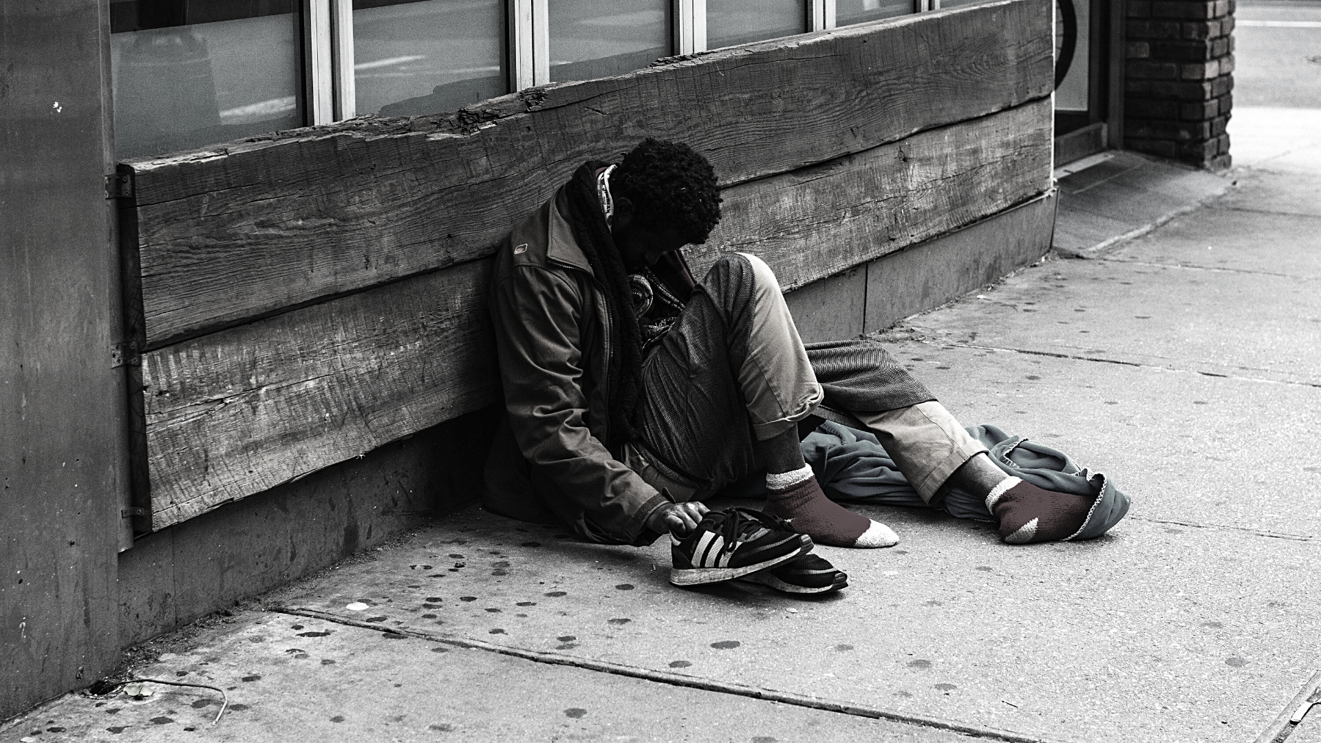 A black man sits on the sidewalk against the side of a building.